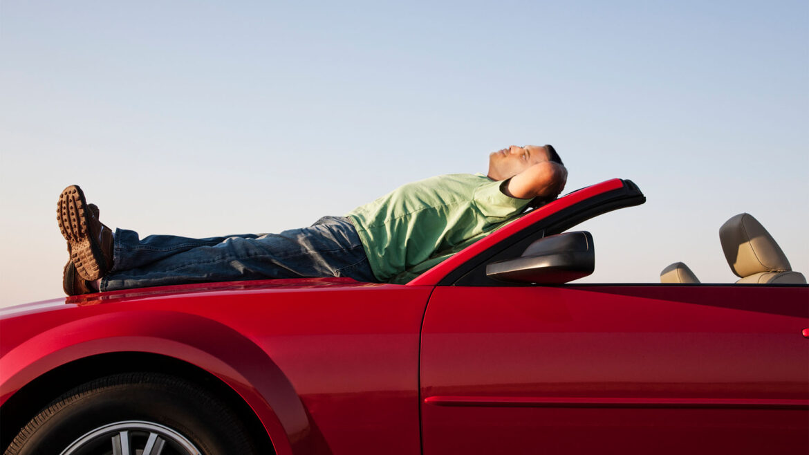 Blue sky background. Young man lying on hood of red convertible, hands behind his head, eyes closed, looking relaxed.