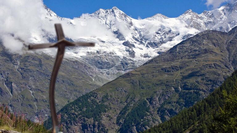 Bent and deformed wooden cross on hillside in isolated mountains with clear blue sky above.