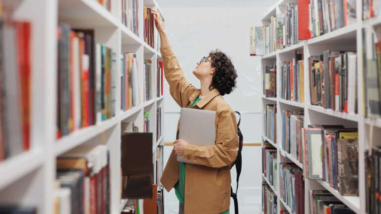 Young woman reaching for a book on a shelf in the library stacks.