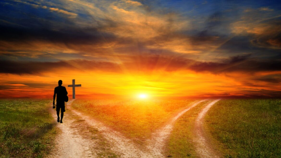 Two dirt roads diverging toward a colorful sunset over a wide open plain. On the left road, a silhouetted single human figure walks towards a cross in the distance.