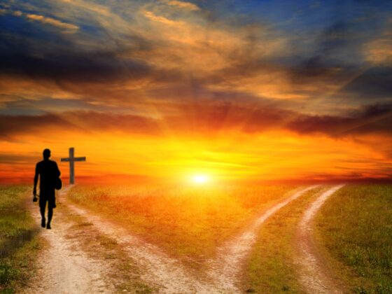 Two dirt roads diverging toward a colorful sunset over a wide open plain. On the left road, a silhouetted single human figure walks towards a cross in the distance.