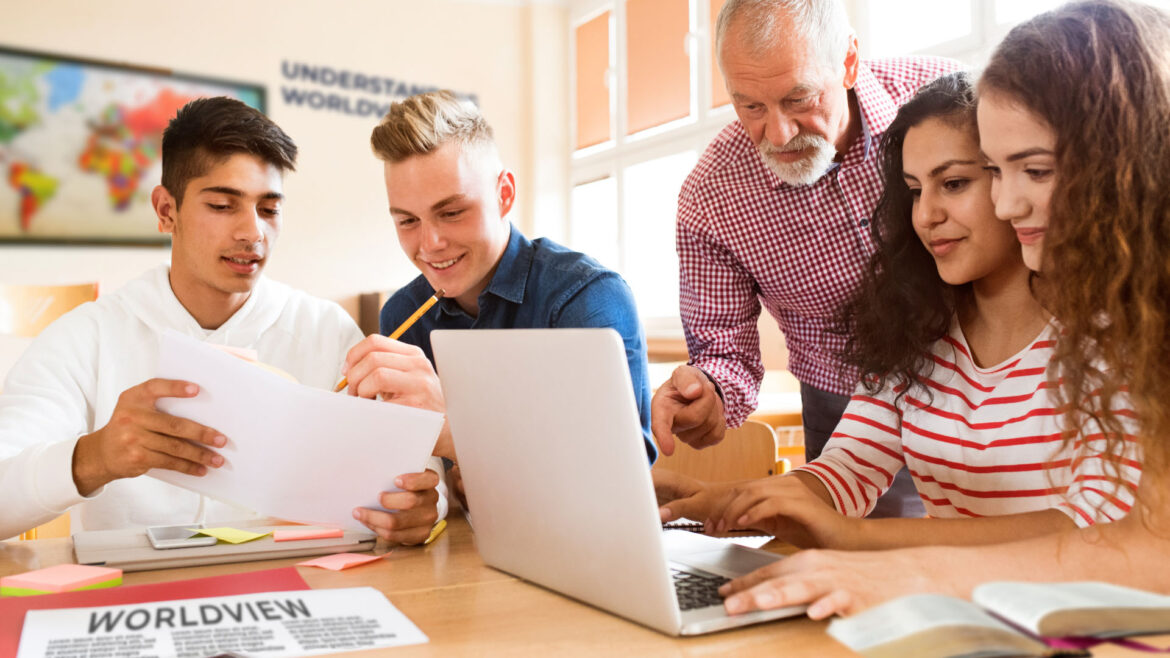 Group of four young adult students and a teacher engaged with print and laptop materials.