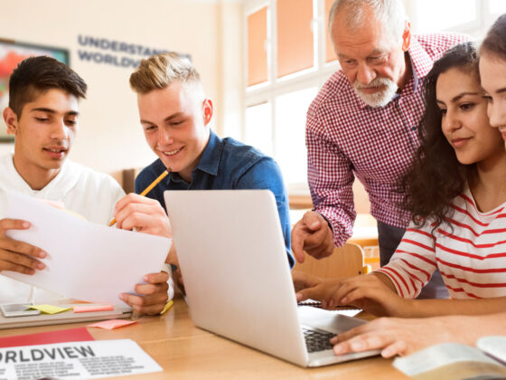 Group of four young adult students and a teacher engaged with print and laptop materials.