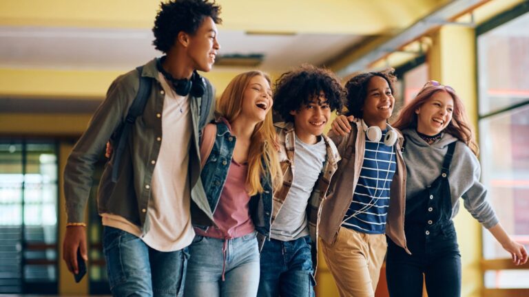 5 young adults with arms around each other walking down a hallway and smiling broadly