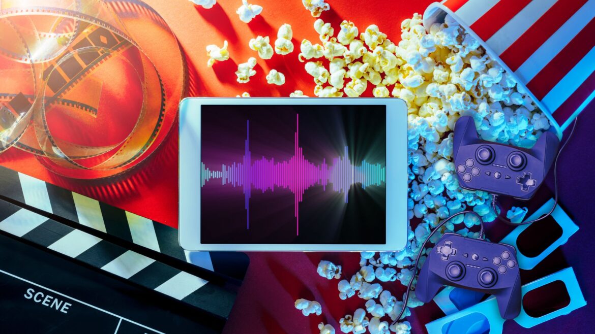 A tablet with colorful digital sound waves laid atop a table with a collection of other entertainment items like popcorn and a move clapper.