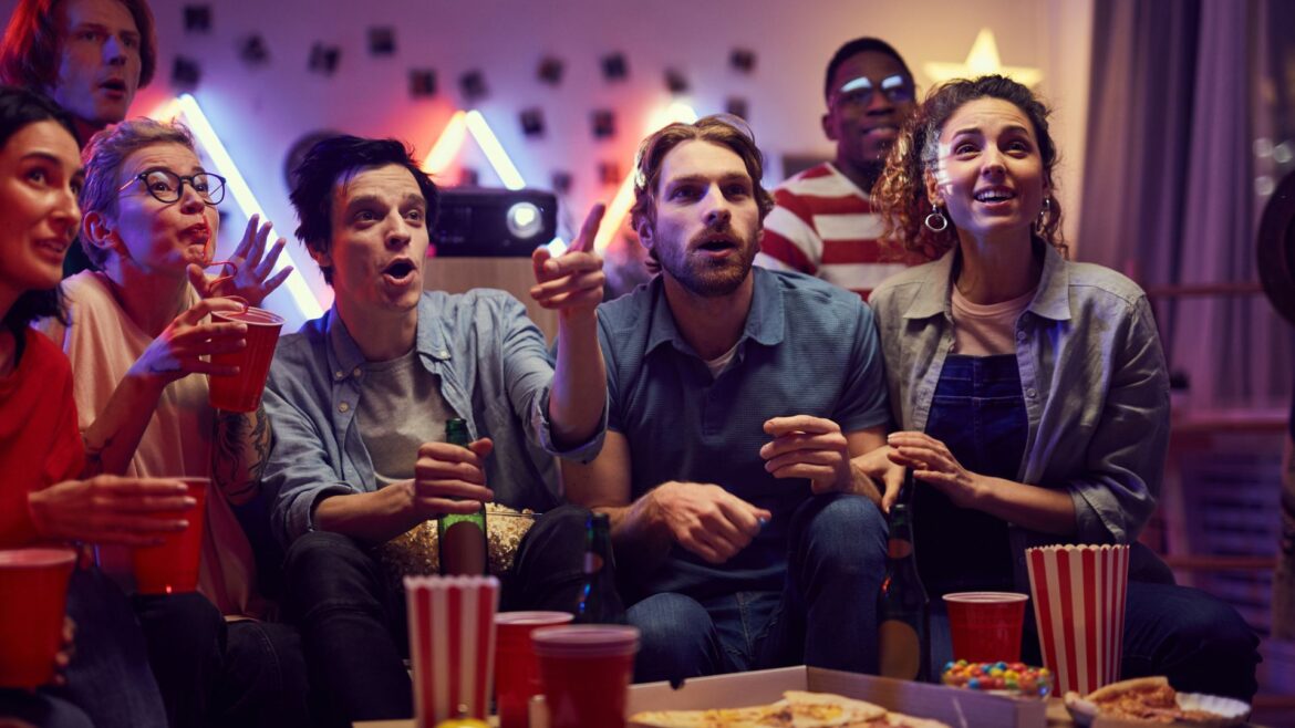 A group of young adults gathered around a low table with snacks as they watch something on a screen out of view in the image.