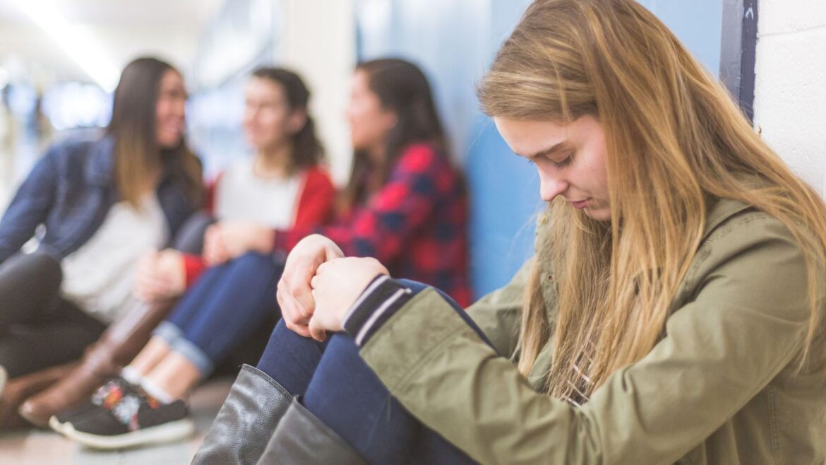 Young adult students seated on the floor in front of a row of lockers. Foreground figure is female with head hung low, hands clasped on knees. Could be praying or just thinking.