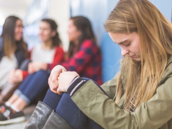 Young adult students seated on the floor in front of a row of lockers. Foreground figure is female with head hung low, hands clasped on knees. Could be praying or just thinking.