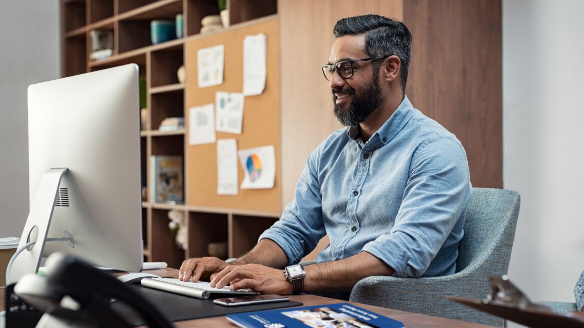 Adult bearded male sitting at a computer keyboard with classroom in background.