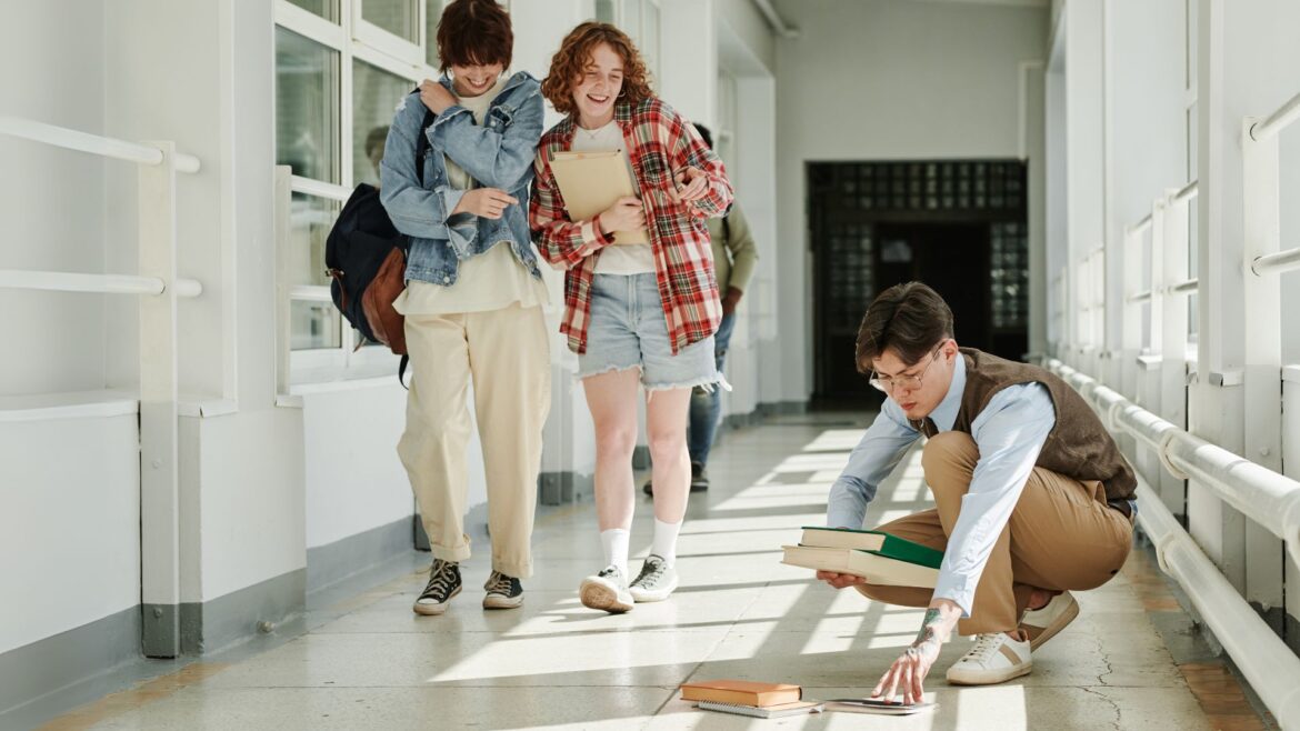 Two students walk by and snicker at a third who has dropped his books on the hallway floor.
