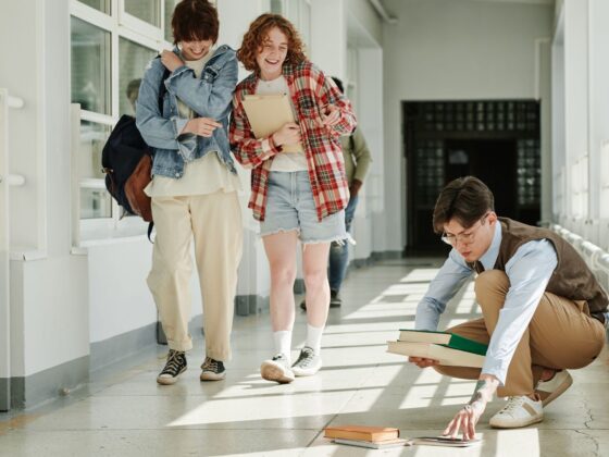 Two students walk by and snicker at a third who has dropped his books on the hallway floor.