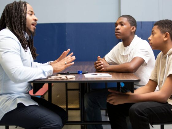 Mentoring conversation between a Christian school educator and two middle or high school students, illustrating spiritual formation, discipleship, and worldview discussion in a school setting.