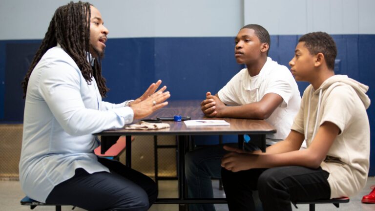 Mentoring conversation between a Christian school educator and two middle or high school students, illustrating spiritual formation, discipleship, and worldview discussion in a school setting.