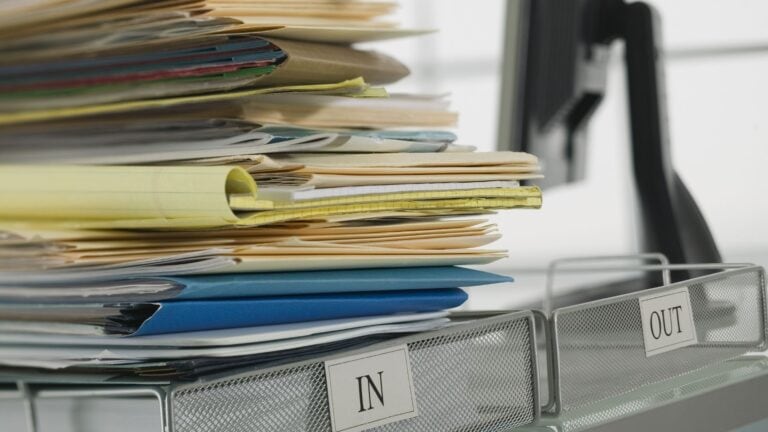 A stack of papers and folders piled high in an office inbox beside an empty outbox.